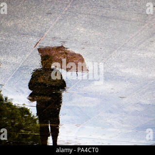 Sfocata riflessione silhouette di una donna che cammina da sola in red ombrelloni , basso angolo di visione, nella pozza in una piovosa giornata di primavera Foto Stock