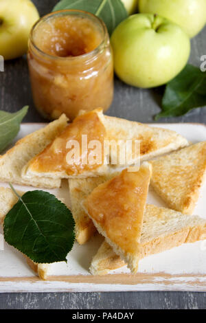 La marmellata di mele in un vasetto di vetro con crostini di pane e mele mature su una superficie di legno. In stile rustico, il fuoco selettivo. Foto Stock