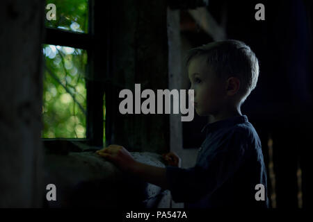 Ragazzo che guarda fuori dalla finestra nella vecchia casa, cercando qualcuno, un paesaggio misterioso, una foto per la copertina del libro. Foto Stock