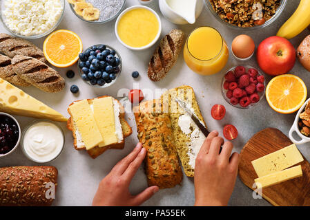 Mani femminili diffusione di burro sul pane. Donna cooking per colazione. Sana colazione ingredienti, telaio di cibo. Muesli, uova, dadi, frutti, bacche, latte, Foto Stock