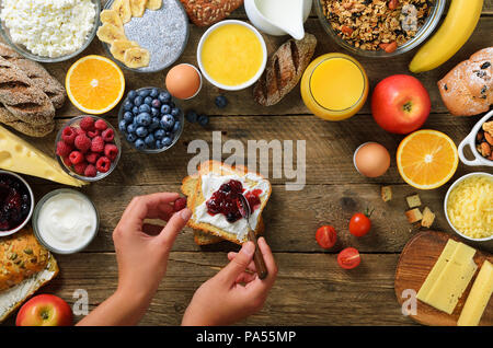 Mani femminili diffusione di burro e marmellata sul pane. Donna cooking per colazione. Sana colazione ingredienti, telaio di cibo. Muesli, dadi, frutti, bacche, mi Foto Stock