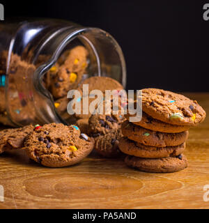 Choc chip cookie smartie cadere al di fuori della jar sul tavolo di legno alto. Foto Stock