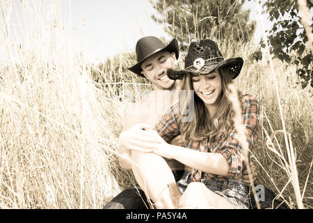 Cowboy e cowgirl giovane tickle reciprocamente durante la seduta in erba lunga indossare cappelli e stivali. Foto Stock