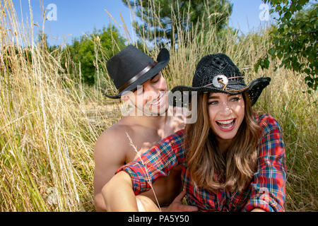 Cowboy e cowgirl giovane tickle reciprocamente durante la seduta in erba lunga indossare cappelli e stivali. Foto Stock