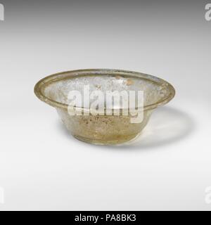 Vaso di vetro. Cultura: Romano. Dimensioni: diametro: 4 3/16 in. × 2 1/16 in. (10,6 × 5,2 cm)Altezza: 1 1/2 in. (3.8 cm). Data: 2a-3a secolo D.C.. Incolore con tingeOutsplayed verdolino, orizzontale bordo arrotondato, ripiegata verso il basso e in su undercurve; lati rastremandosi verso il basso; anello di base; profonda, fondo concavo con cicatrice pontil.intatti; pinprick bolle; dulling, vaiolature e iridescenza, con piccole macchie di resistenza agli agenti atmosferici di consistenza cremosa. Museo: Metropolitan Museum of Art di New York, Stati Uniti d'America. Foto Stock