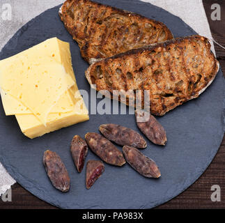 Pezzi di salame affumicato, formaggio e pane tostato dal pane fatto con farina di grano su una lastra di grafite, vista dall'alto Foto Stock