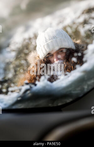 Bella bruna donna pulizia vetro auto da neve e ghiaccio. La raschiatura di ghiaccio da un parabrezza è stato preso dall'interno dell'auto. Trasporto Foto Stock