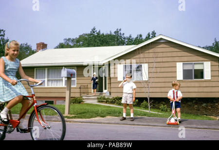 Famiglia americana che indossa abiti domenicali comprendenti madre e tre bambini piccoli fuori casa di recente costruzione, Briarcliff Manor, Westchester County, New York, USA nel 1963. La ragazza bionda sta guidando una bici ed i ragazzi giovani stanno portando gli shorts. Il bambino sta giocando con un tosaerba giocattolo Foto Stock