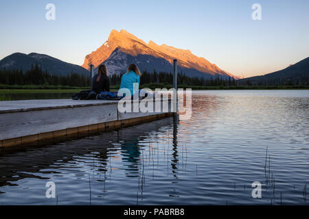 Giovane amici stanno godendo della splendida estate tramonto su un dock in legno dall'acqua. Preso in laghi Vermiglio, Banff, Alberta, Canada. Foto Stock