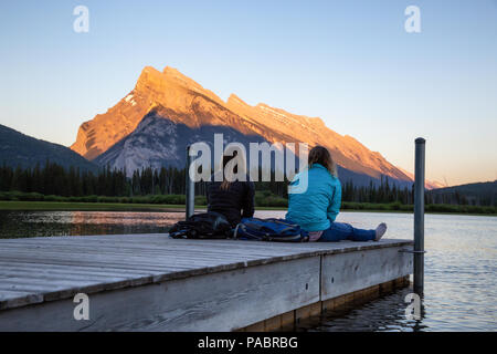 Giovane amici stanno godendo della splendida estate tramonto su un dock in legno dall'acqua. Preso in laghi Vermiglio, Banff, Alberta, Canada. Foto Stock