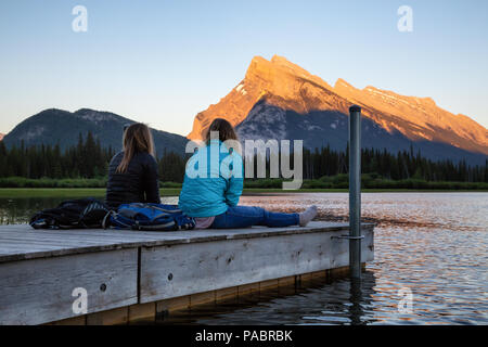 Giovane amici stanno godendo della splendida estate tramonto su un dock in legno dall'acqua. Preso in laghi Vermiglio, Banff, Alberta, Canada. Foto Stock