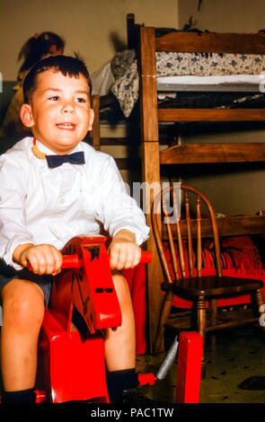 Ragazzo sorridente che indossa una camicia bianca e una cravatta ad arco cavalcare un giocattolo di legno a cavallo in una camera da letto per bambini Con letti a castello negli Stati Uniti negli anni '60 Foto Stock