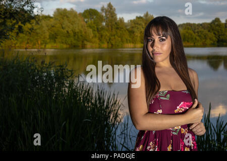 Lo spagnolo donna americana in piedi sul bordo di un lago in Cambridgeshire. Indossando una susina colorato abito floreale durante un tramonto d'estate. Foto Stock