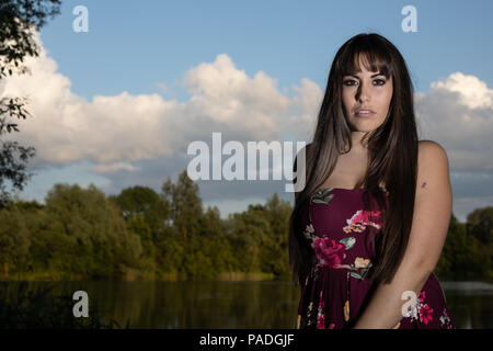 Lo spagnolo donna americana in piedi sul bordo di un lago in Cambridgeshire. Indossando una susina colorato abito floreale durante un tramonto d'estate. Foto Stock