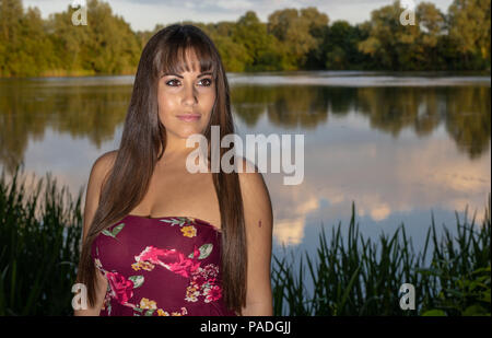 Lo spagnolo donna americana in piedi sul bordo di un lago in Cambridgeshire. Indossando una susina colorato abito floreale durante un tramonto d'estate. Foto Stock