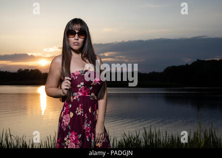 Lo spagnolo donna americana in piedi sul bordo di un lago in Cambridgeshire. Indossando una susina colorati abiti floreali e occhiali da sole durante un tramonto d'estate. Foto Stock