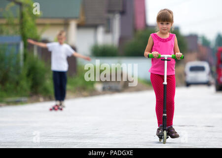 Piuttosto giovane dai lunghi capelli biondi ragazza bambino in rosa vestiti con gli scooter sul sobborgo di sole street e ragazzo su skateboard sfocato estate sfondo luminoso. Foto Stock