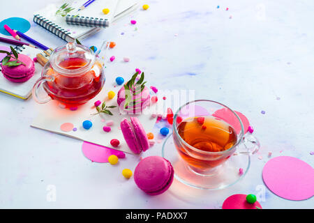 Teiera vetro testata con caramelle e coriandoli su uno sfondo luminoso con copia spazio. Rosa e viola tavolozza ancora in vita. La vibrante tea party drink concept Foto Stock
