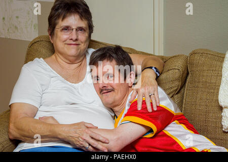 Un vecchio uomo con la sindrome di Down, abbracci la sua sorella come essi sedersi su un divano. La donna guarda stanco, l'uomo guarda mischeivious. Egli è in procinto di tic Foto Stock