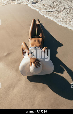 Donna in bikini a prendere il sole sdraiati vicino al mare indossando cappello per il sole con le onde del mare arrivano fino a lei. La donna in vacanza indossando cappello per il sole di relax presso il beac Foto Stock