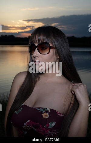 Lo spagnolo donna americana in piedi sul bordo di un lago in Cambridgeshire. Indossando una susina colorati abiti floreali e occhiali da sole durante un tramonto d'estate. Foto Stock