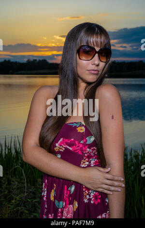 Lo spagnolo donna americana in piedi sul bordo di un lago in Cambridgeshire. Indossando una susina colorati abiti floreali e occhiali da sole durante un tramonto d'estate. Foto Stock