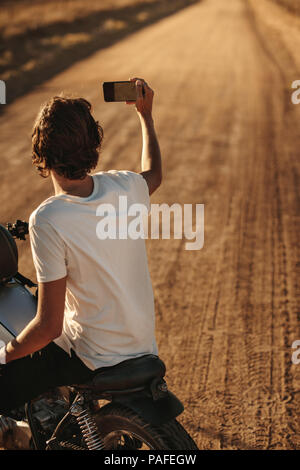 Vista posteriore del giovane uomo prendendo selfie con il suo smart phone mentre è seduto sulla sua moto sulla strada di campagna. Bike Rider catturare i ricordi del suo countr Foto Stock