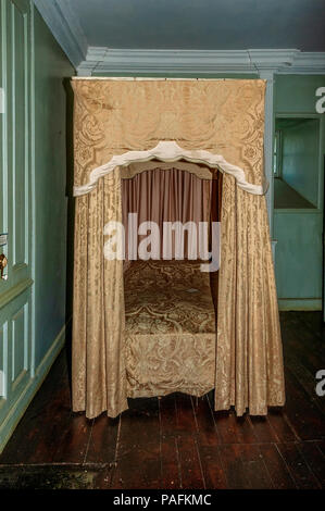 Un letto a baldacchino con tende a motivi geometrici poggia su un pavimento di legno vicino al dipinto di pareti in pannelli di legno vicino ad una finestra porta pistola Foto Stock