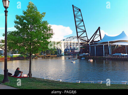 Un giovane si siede nel greenspace sulla riva est del Cleveland Ohio appartamenti affacciato sul fiume Cuyahoga e Jacobs Padiglione sul West Bank. Foto Stock