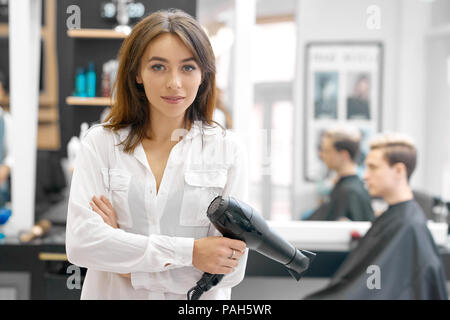 Apparecchio femmina mantenendo potente asciugacapelli guardando la telecamera, in posa spacy beaty salon. Giovane maschio cliente seduto vicino a un grande specchio sullo sfondo. Donna che indossa classico bianco shirt, sorridente. Foto Stock