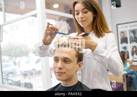 Parrucchiere femmina facendo del taglio di capelli per il bel modello con tonico lilla. Parrucchiere utilizzando metallici affilati e forbici plestic nera pettine. Master che indossa una camicia bianca e guardando la fotocamera. Foto Stock