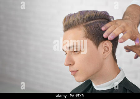 Giovane maschio modello in attesa di nuova acconciatura di lilla la colorazione dei capelli. Parrucchiere mani facendo elegante colorazione per bel ragazzo. Lavorando con mattoni bianchi di sfondo per studio. Foto Stock