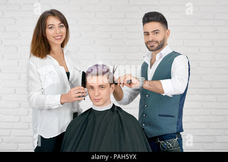 Maschi e femmine di parrucchieri facendo del taglio di capelli per giovani client con capelli tonico. Utilizzando pettini in plastica per lo styling e taglienti forbici metallico. Barbiere indossando gilet, donna avente classico bianco shirt. Foto Stock