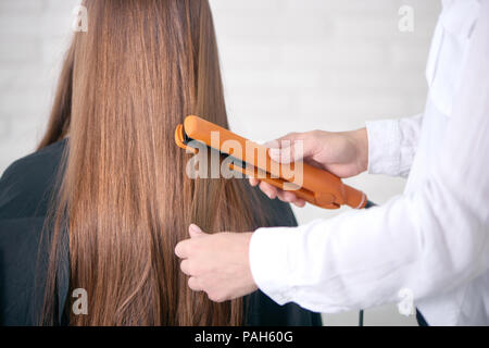 Vista posteriore del parrucchiere straighting client di capelli lunghi marrone. Modello avente liscia e diritta, glancy, capelli sani, seduta ricoperta con speciali capo nero. Apparecchio indossando classico bianco shirt. Foto Stock