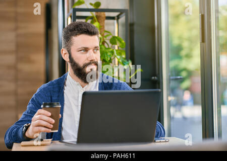 Attraente uomo con la barba e gli occhi blu a lavorare con il computer portatile in prossimità di grandi finestre panoramiche in ristorante con interni eleganti. Avendo il marrone scuro capelli blu brillante cardigan e camicia bianca. Bere caffè Foto Stock