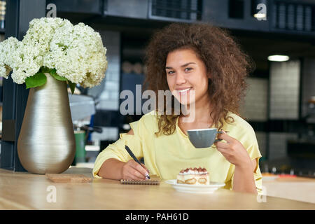 Sorridente ragazza con i capelli ricci di bere il caffè, la scrittura di note durante la seduta in un ristorante moderno. Deliziosi dolci della pace di torta in piedi sul tavolo di legno vicino al vaso metallico bianco con fiori lilla. Foto Stock