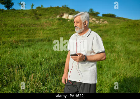 Il vecchio uomo mantenendo il telefono cellulare, in piedi sul campo guardando a lato. Ascolto di musica con cuffie rosso. Indossando il bianco classico Polo shirt scuro con strisce blu,pantaloni neri, sneakers. Stile di vita sportiva. Foto Stock