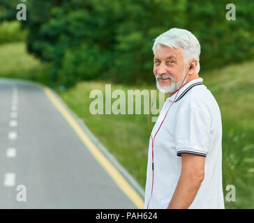 Sorridente uomo vecchio permanente sulla città racetrack guardando la fotocamera. Indossando il bianco classico Polo shirt scuro con strisce blu, sport watch, red headphones. Sport all'aperto, corsi di formazione. Foto Stock