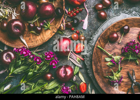 Estate frutta di stagione e frutti di bosco con fiori da giardino in piastre su scuro dello sfondo rustico, vista dall'alto Foto Stock