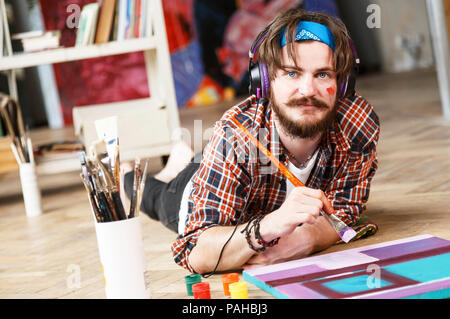 Bello dai capelli scuri tanga barbuto artista maschile nelle cuffie e blu bandana giace sul pavimento e la spazzola di contenimento nel suo studio contemporanea con molti dipinti Foto Stock