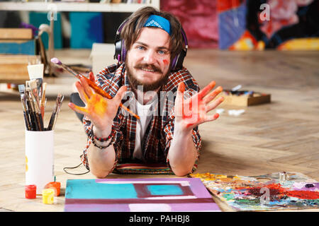 Bello dai capelli scuri tanga barbuto artista maschile nelle cuffie e blu bandana giace sul pavimento e mostra le mani nel suo studio contemporanea con molti dipinti Foto Stock