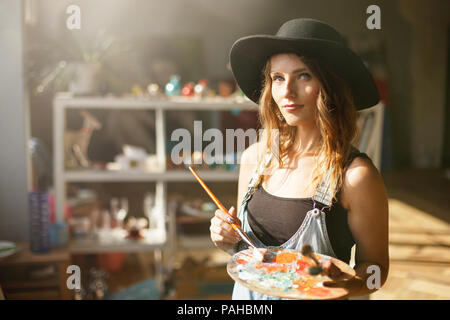 Ritratto di bello a lungo i capelli artista femminile nella cute black hat di miscelazione dei colori della tavolozza e schizzi in contemporanea studio con molti dipinti Foto Stock