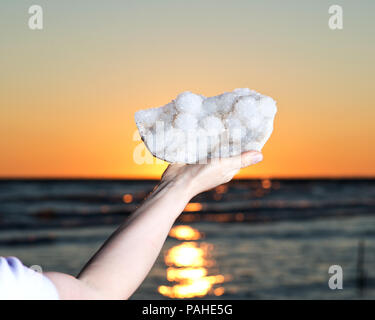 Donna che mantiene Aurora Rainbow cluster di quarzo da India nella sua mano al sorgere di fronte al lago. Foto Stock