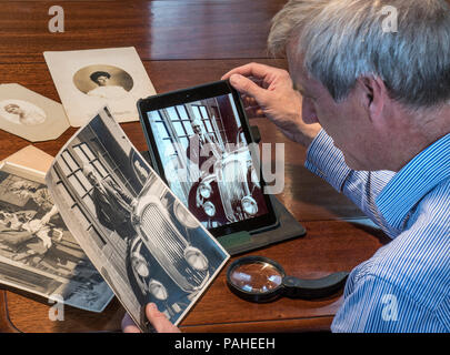 Uomo maturo ordinamento tramite le vecchie fotografie confronta un originale B&W stampa con la sua immagine scansionata sul suo computer tablet Foto Stock