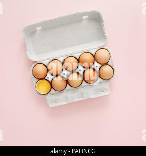 Uovo di pollo è mezzo rotto tra le altre uova in cartone uovo su sfondo rosa vista superiore, laici piatta Foto Stock