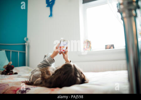 Bambina è sdraiato sul suo letto, la riproduzione di un gioco sul suo smartphone. Foto Stock