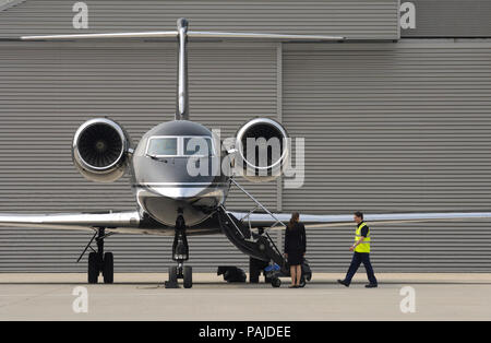 Un uomo che indossa un tabard giallo e una hostess in piedi dal airstairs di aviazione Gama Gulfstream 4SP parcheggiato con il tag hangar dietro Foto Stock