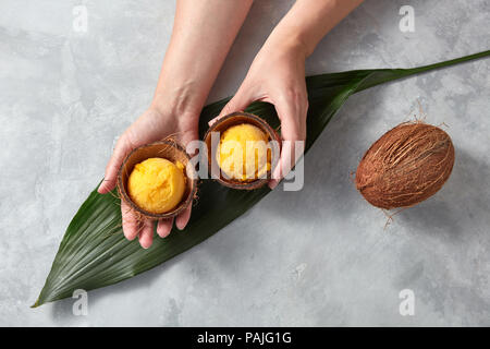 Mani femminili tenere due metà di guscio di noce di cocco con palline di Giallo mango gelato sopra il verde di piante tropicali e foglia tutta una noce di cocco su un marmo grigio Sfondo. Vista dall'alto. Foto Stock