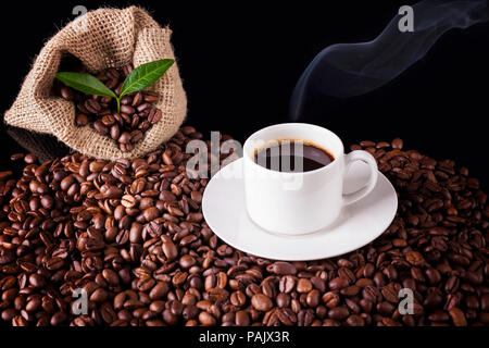 Caffè fumante accanto a un sacco di tela riempito con i chicchi di caffè, collocato su un mucchio di chicchi di caffè, su sfondo nero Foto Stock