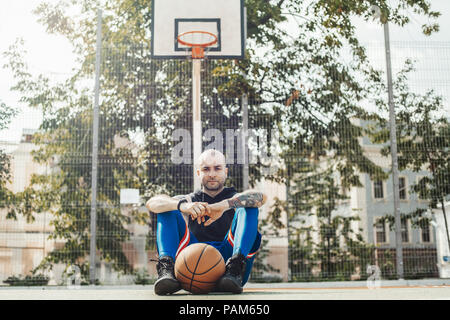 Bald attraente uomo seduto sotto canestro da basket e la scheda sul campo da basket. L uomo è sul fuoco e primo piano, Basketball hoop e la scheda su Foto Stock